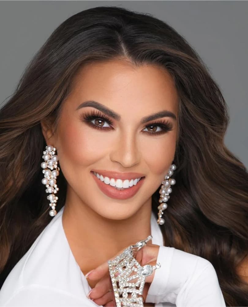 Elegant woman in a white outfit holding a sparkling crown, smiling confidently with long wavy hair and pearl earrings, representing Miss Miami Pageants.