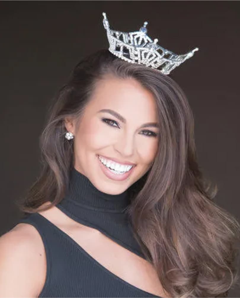 Miss Miami titleholder wearing a crown and a black evening dress, smiling confidently against a dark background.