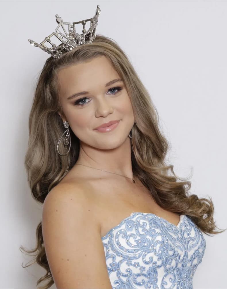 Teen pageant contestant in a strapless light blue gown with curled hair, smiling softly in front of a white backdrop.
